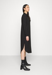 Vestido preto de manga longa com fecho de botão e fendas laterais, feito de tecido suave. Acompanhado de botas altas pretas com bico quadrado.