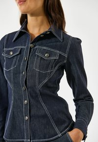 Ciemnoniebieska koszula dżinsowa o dopasowanym fasonie, z białym przeszyciem, dwoma kieszeniami na piersi i metalowymi guzikami, podkreślona spiczastym kołnierzykiem.
