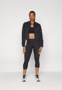 Svart kort jakke over en svart treningscrop-top, paret med svarte capri leggings og hvite joggesko. Glatt stoff, tettsittende design.