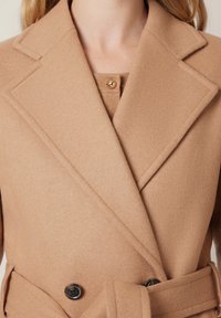 Płaszcz z mieszanki wełny w odcieniu kamelowym, z wyciętym kołnierzem, podwójnym zapięciem i wiązanym paskiem. Pod spodem znajduje się zapinany na guziki sweter.