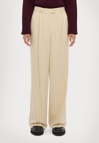 Beige, gestreifte weite Hosen mit einem taillierten Bund und Falten. Der Stoff wirkt weich und ergänzt schwarze klobige Schuhe.