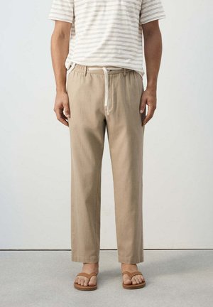 Hombre con pantalones beige con cordón, una camiseta de manga corta a rayas blancas y marrones, y sandalias marrones, de pie sobre un suelo claro.