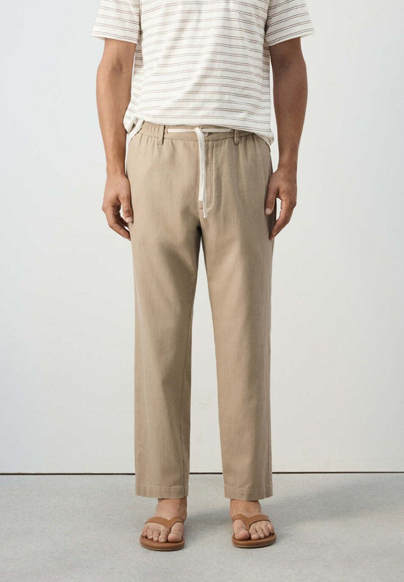 Homme portant un pantalon beige à cordon, une chemise à manches courtes rayée blanche et marron, et des sandales marron, debout sur un sol clair.