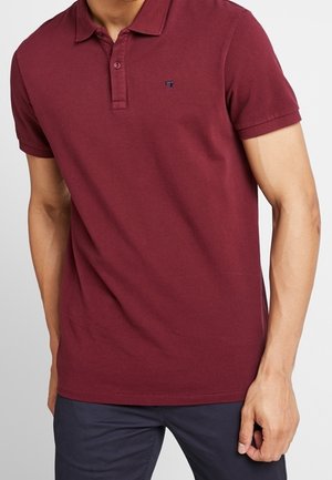 Uomo che indossa una polo aderente a maniche corte di colore rosso scuro con colletto e bottoni, abbinata a pantaloni blu scuro.