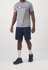 Szara koszulka z krótkim rękawem z logo "New Balance", zestawiona z granatowymi szortami i ciemnymi sneakerami, z fakturowaną podeszwą i białymi skarpetkami.