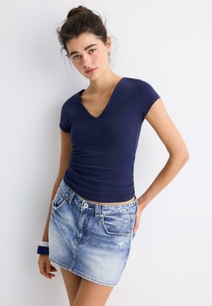Giovane donna con capelli scuri raccolti in uno chignon, che indossa una maglietta blu navy con scollo a V e una minigonna di jeans azzurro chiaro su sfondo bianco.