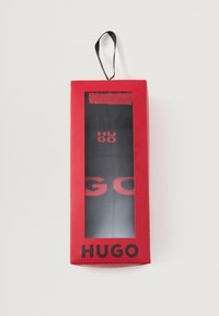 Čierne ponožky s červeným brandingom "HUGO" v transparentnej výstavnej krabičke, s červeným vonkajším obalom a visačkou pre jednoduché vystavenie.