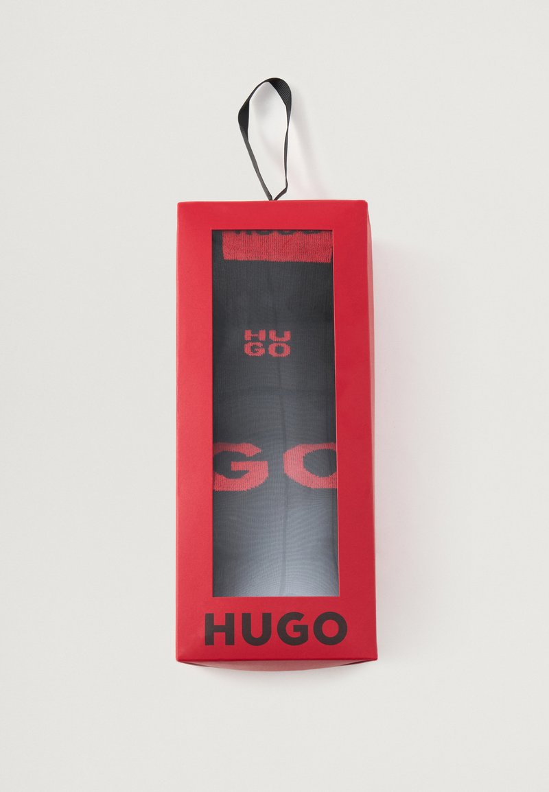 Čierne ponožky s červeným brandingom "HUGO" v transparentnej výstavnej krabičke, s červeným vonkajším obalom a visačkou pre jednoduché vystavenie.