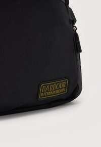 Γωνία μαύρης υφασμάτινης τσάντας με κίτρινη και μαύρη ετικέτα λογοτύπου Barbour International και ορατό πλαϊνό φερμουάρ με τανάλια.