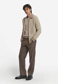 Beige ribbad stickad jacka med krage, buren över en ljusbeige tröja, kombinerad med skräddarsydda bruna byxor och svarta skor.