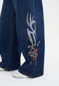 Ruudukankaiset siniharmaat farkut, joissa on painatettuja kuvioita: sydän, jossa on "PARIISI" ja eloisat, abstraktit elementit sivuilla.