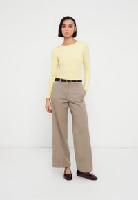 Φωτεινό κίτρινο μακρυμάνικο τοπ, μπεζ παντελόνι με πλατιά μπατζάκια και ψηλή μέση, μαύρη ζώνη και καφέ σκούρα λουστρινένια μοκασίνια. Απλό φόντο.