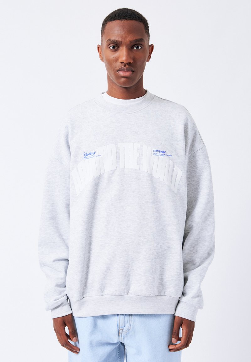 Sudadera gris claro con cuello redondo, con un corte oversize y el texto "AROUND THE WORLD" en blanco. Textura suave y puños acanalados.
