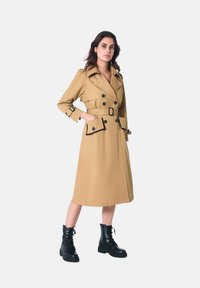 Manteau trench kaki à double boutonnage, accents noirs, grands boutons et taille ceinturée, associé à des bottes de combat noires à lacets.