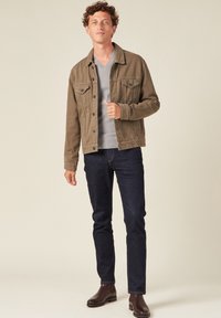 Giacca in denim cotone marrone con tasche a bottoni, maglione grigio chiaro a scollo a V, jeans blu scuro e stivali alla caviglia in pelle marrone.