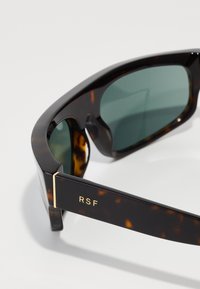 Skilpadde-solbriller med mørk grøn linser, med et slankt design og guldakcent med "RSF" merking på siden.