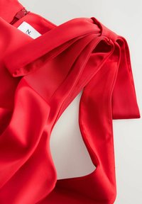 Červený saténový top s mašlí na rameni, má hladkou strukturu a volný střih. Na zádech je viditelný zip.