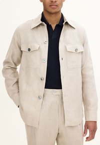 Beige linned skjortejakke med knaplukning, to brystlommer og flad krave, båret over en marineblå mønstret strikpolo.