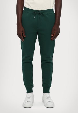 Uomo che indossa pantaloni da jogging verde scuro con vita a coulisse e scarpe da ginnastica bianche, in piedi davanti a uno sfondo bianco semplice.