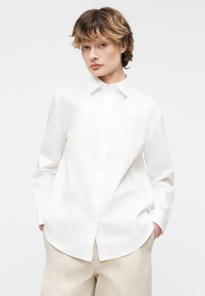 Άτομο με κοντά κυματιστά μαλλιά, φοράει άσπρο πουκάμισο με κουμπιά και μπεζ παντελόνι, στέκεται με τα χέρια στις τσέπες μπροστά σε απλό φόντο.