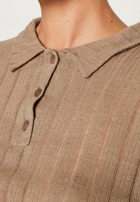 Ruskea ribattukuvioinen polo-paita, jossa on teräväkaulus ja kolme nappia. Kankaassa on pehmeä tekstuuri ja hillityt pystysuorat raidat.