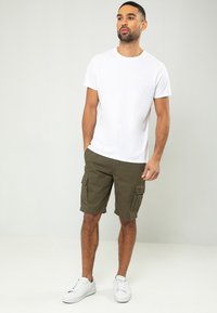 T-shirt blanche à col rond associée à un short cargo vert olive avec des poches latérales, portée avec des baskets blanches sur un fond uni.