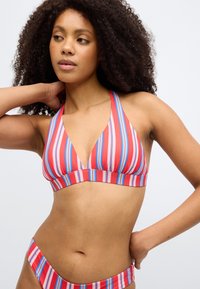 Svītrainu bikini tops sarkanā, baltā un zilā krāsā; trijstūrveida kausi ar platu joslu. Audums izskatās gluds ar regulējamām siksnām. Modele pozē, viena roka aiz kakla.