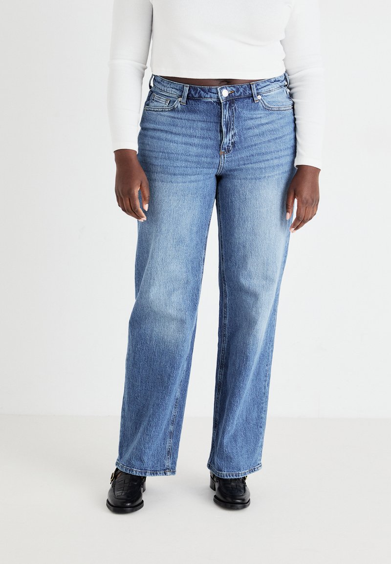 Lys blå, høyt liv jeans med avslappet passform, med fem lommer og knappelukking. Brukt med en hvit langermet topp og svarte sko.