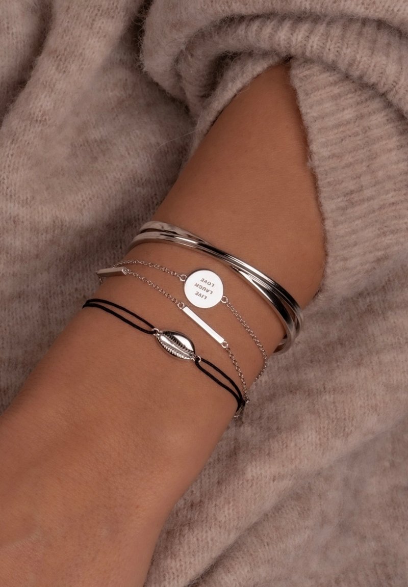 Trys apyrankės ant riešo: dvi sidabrinės bangles, viena sidabrinė grandinėlė su apvaliu pakabuku ir viena juoda virvutės apyrankė su kriauklės detale.