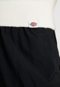 Black fabric pants with a textured finish, featuring a white ribbed waistband and a Dickies logo tag on the side.