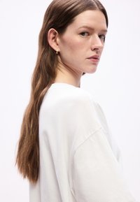 Νέα γυναίκα με μακριά καστανά μαλλιά, φοράει λευκό πουκάμ ισο και μικρά χρυσά κρίκους στα αυτιά, κοιτάζοντας πάνω από τον ώμο της σε απλό φόντο.