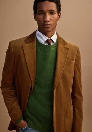 Man wearing brown corduroy blazer, green sweater, striped shirt, and brown tie against beige background, hand in pocket.