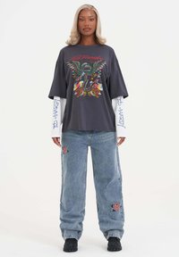 Grijze oversized T-shirt met kleurrijke draakafbeelding en tekst, gelaagd over een wit hemd met lange mouwen. Lose blauwe jeans met rozenborduursel.