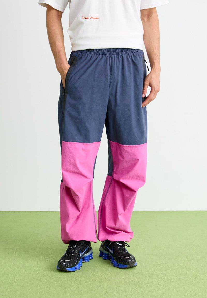 Personne portant un pantalon coloré en blocs de bleu et de rose avec des baskets noires et bleues, se tenant sur un sol vert devant un fond blanc.