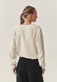 Weiße Bluse mit langen Ärmeln, weicher Textur und lockerer Passform, mit Rundhalsausschnitt. Tragen Sie sie mit schwarzen, taillierten Hosen.