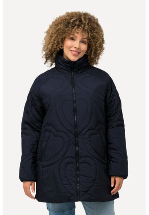 Marineblå quilted puffer-jakke med høj krave, hjerteformede quilted detaljer, lynlås lukning og sidelommer, iført over lyse denimbukser.