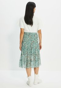 Jupe en tissu floral à volants de couleur verte avec un matériau fluide et transparent. Elle présente de petites fleurs blanches et orange ainsi qu'une taille élastique. Bottines blanches jusqu'aux chevilles.