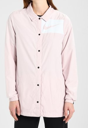 Personne portant une veste boutonnée rose clair Nike avec un col et des poignets élastiques, avec un panneau blanc et un logo Nike swoosh estompé sur la poitrine.
