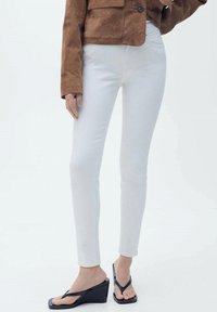 Bele skinny jeans z gladko teksturo, kombinirane z rjavo zamšasto jakno. Model nosi črne klinaste sandale z edinstveno zasnovo.