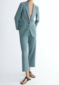 Personne portant un tailleur-pantalon bleu sarcelle avec un pantalon droit court et des sandales noires à lanières, debout les mains derrière le dos.