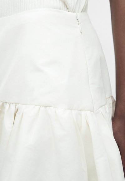 Gros plan d'une jupe blanche taille haute avec un ourlet froncé et une fermeture éclair latérale, portée avec un top côtelé crème.