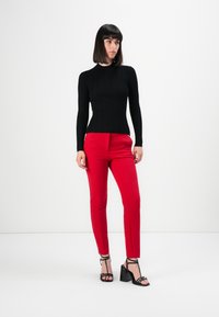 Žena stojící proti bílému pozadí, oblečená v černém žebrovaném topu s dlouhým rukávem, červených slim-fit kalhotách a černých sandálech na vysokém podpatku.