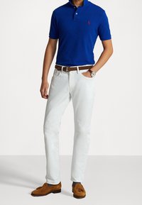 Blå pikétröja med krage och logotyp, vita jeans, brun läderbälte och bruna mockaskor. Enkel, avslappnad look utan mönster.