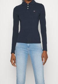 Person in einem taillierten navyblauen Langarm-Polohemd mit drei Knöpfen und hellblauen Jeans vor einem weißen Hintergrund.