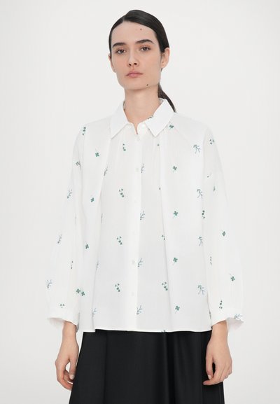 Chemise blanche à boutons avec broderie de feuilles vertes, manches longues bouffantes et col. Tissu doux et léger avec une coupe décontractée.