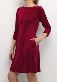Röd sammetklänning med lång ärm, rundad halsringning, sidofickor och samlad textur på ärmarna. Faller ovanför knäet.