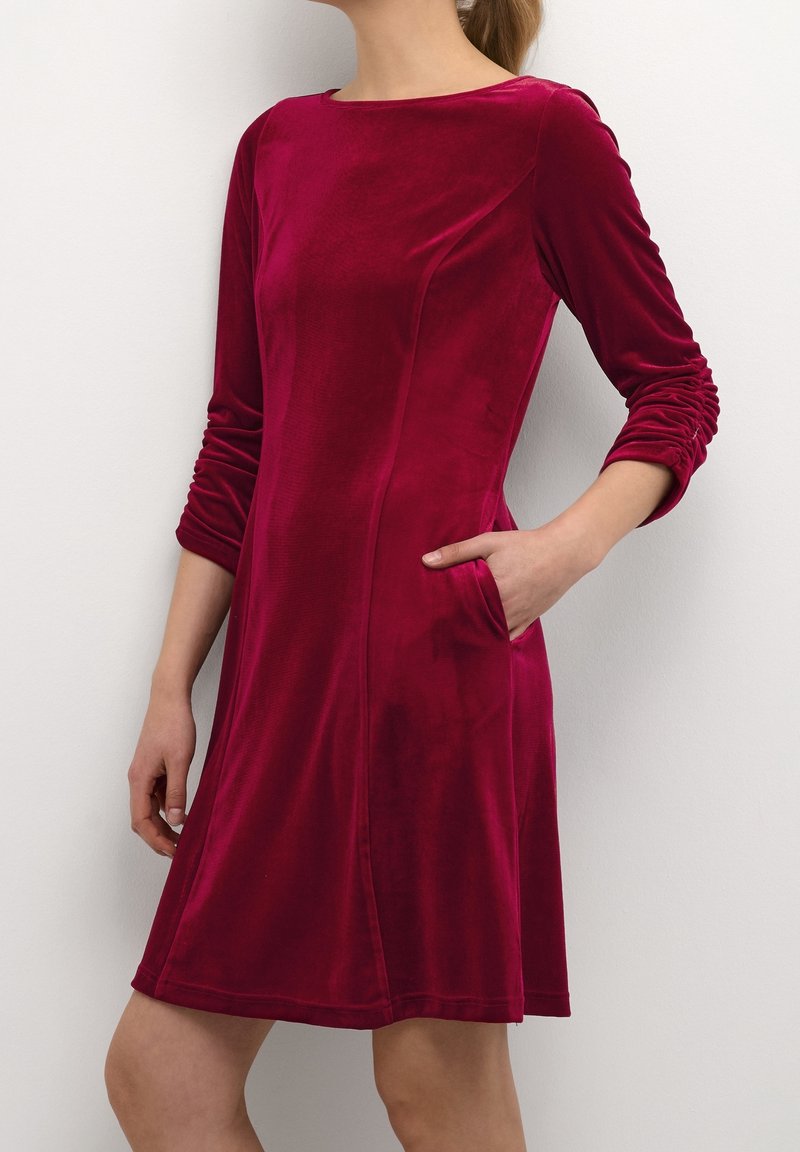Röd sammetklänning med lång ärm, rundad halsringning, sidofickor och samlad textur på ärmarna. Faller ovanför knäet.