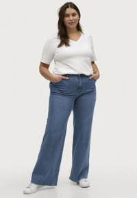Lettet, hvid kortærmet V-hals t-shirt parret med højtaljede, brede blå jeans. Modellen har hvide sneakers på; hænderne i lommerne.