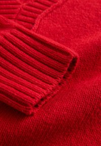 Red knitted sweater with ribbed cuffs and textured body. Smooth and soft fabric, showing a close-up of the stitching details.