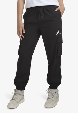 Person iført sorte cargo joggers med elastiske manchetter og hvide sneakers, hænder i lommerne, viser Jumpman-logoet på bukserne.